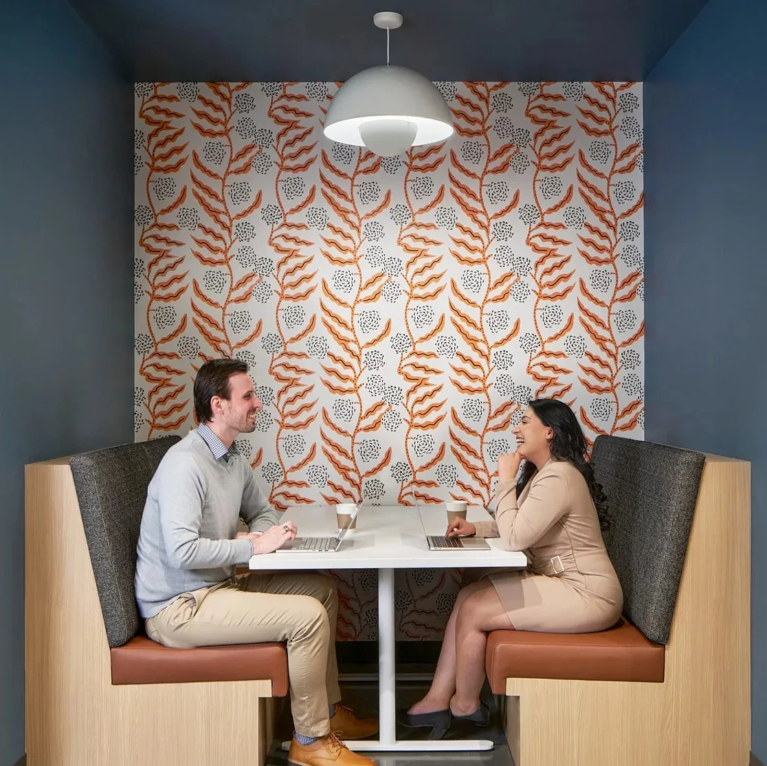 Two coworkers chatting in a office booth with botanical wallpaper on the wall