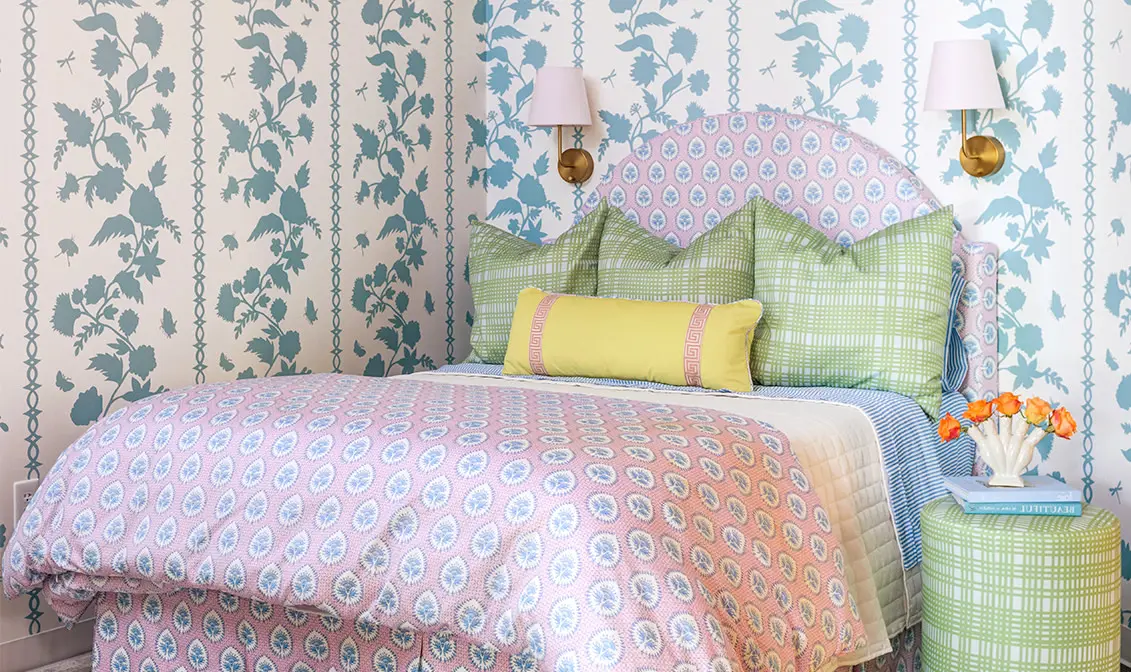 Pink and green bedding with matching upholstered headboard in room with blue wallpaper.